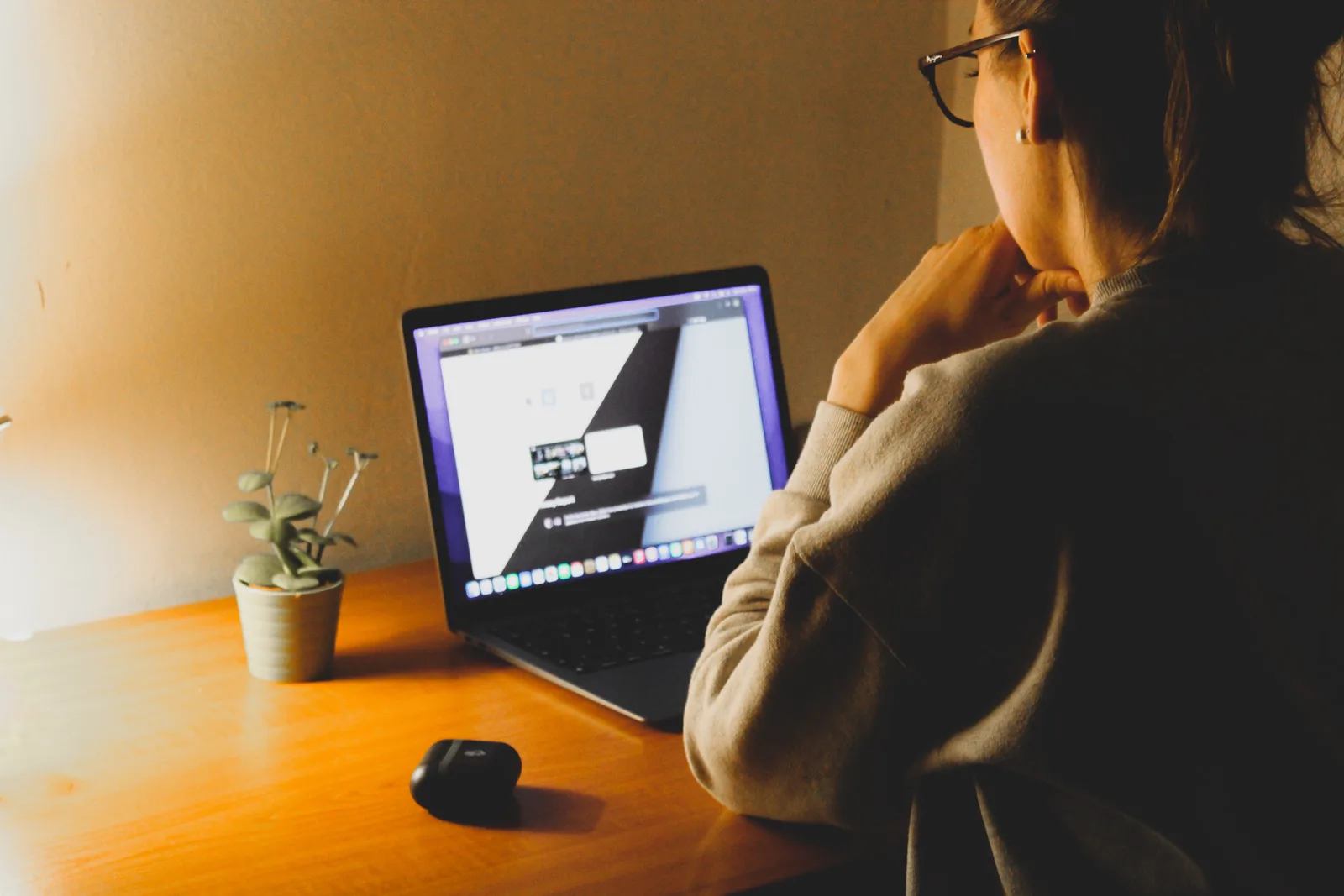 A woman working on a laptop at a wooden desk with a plant under warm lighting.