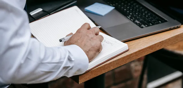 Close-up of an adult writing in a notebook beside a laptop. Professional setting. Audience research for a website project that keeps buyers and end users apart