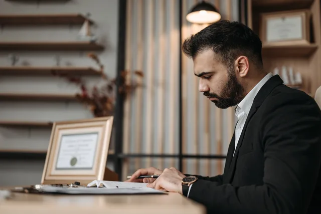 A professionally dressed man focused on writing at a desk within an office setting, suggesting a serious work environment. Commercial contracts for web design agencies: seven records that keep a commission honest